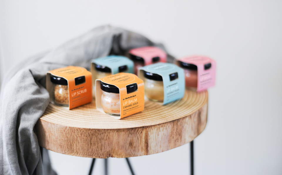 Photo of six lip balms on a wooden table in small jars with colourful paper sleeves as labels.