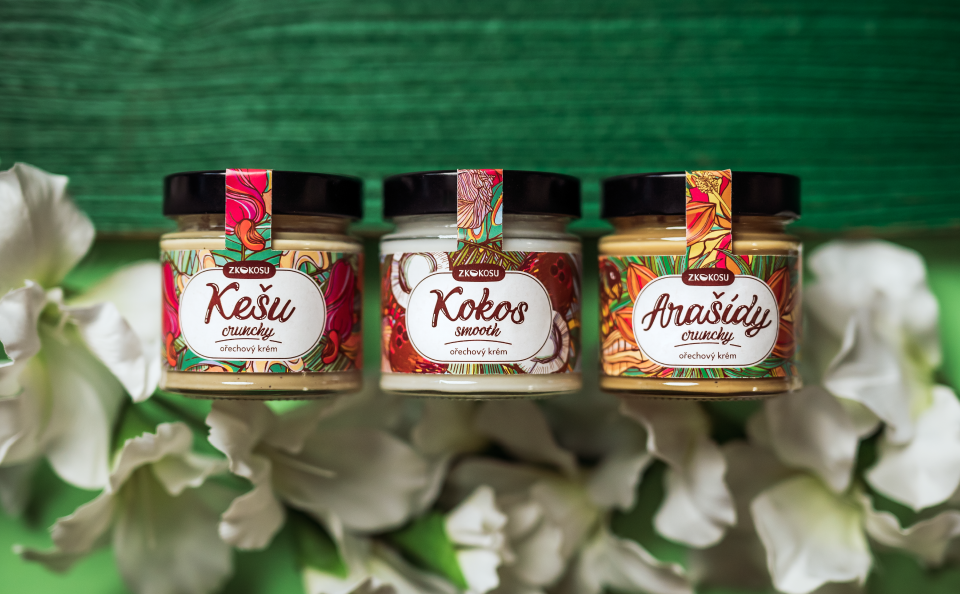 Photo of nut creams made from cashew nuts, coconut and peanuts in a glass jar, on the illustrated labels there are tropical motifs referring to the main ingredients in the composition. In the background are palm leaves and white flowers.