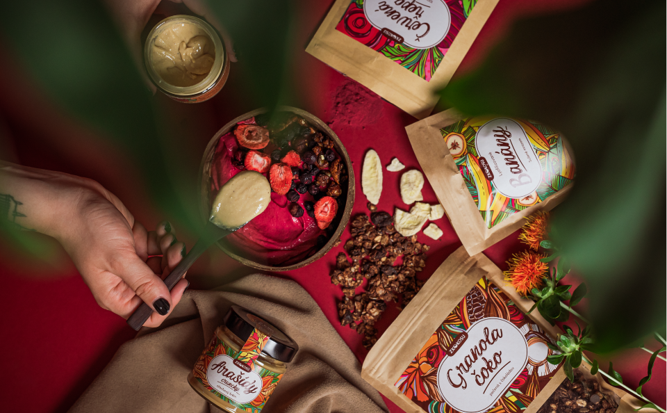 Mood photo of food products from the e-shop zkokosu.cz - peanut butter, dried bananas, beetroot superfood powder and chocolate granola. A woman's hand holds a wooden spoon with peanut butter on it, in the background there is a red smoothie with dried fruit and granola topping in a coconut bowl. Photographed in low light on a red background.