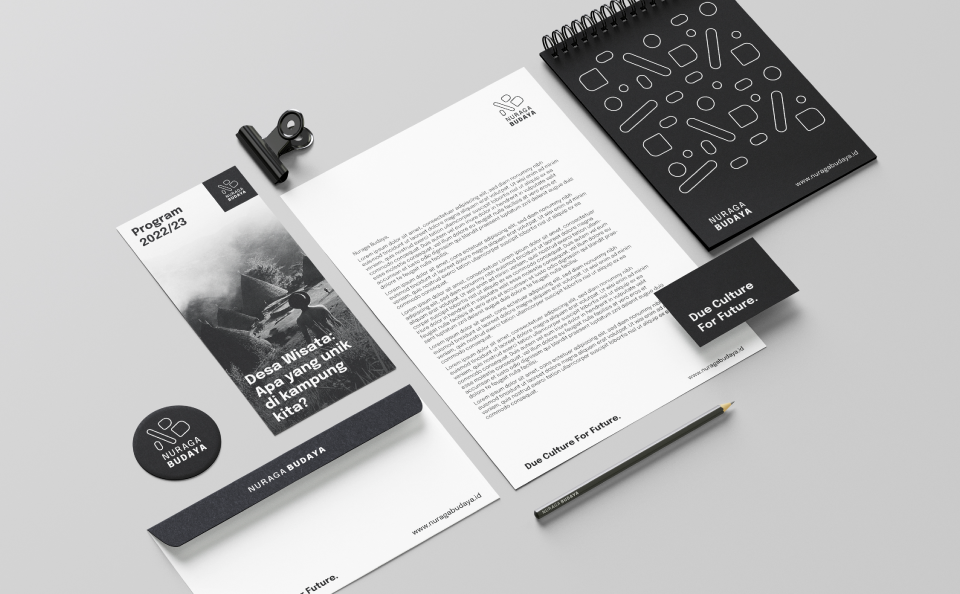 Visualization of Nuraga Budaya stationary in basic black and white colour on a gray background. The following items are placed diagonally on the picture: a black round sticker with a white logo, a 2022-2023 webinar program flyer, a DL envelope, letter paper, a business card, an A5 notepad and a black pencil. The items have either the logo or the claim "Due Culture For Future"