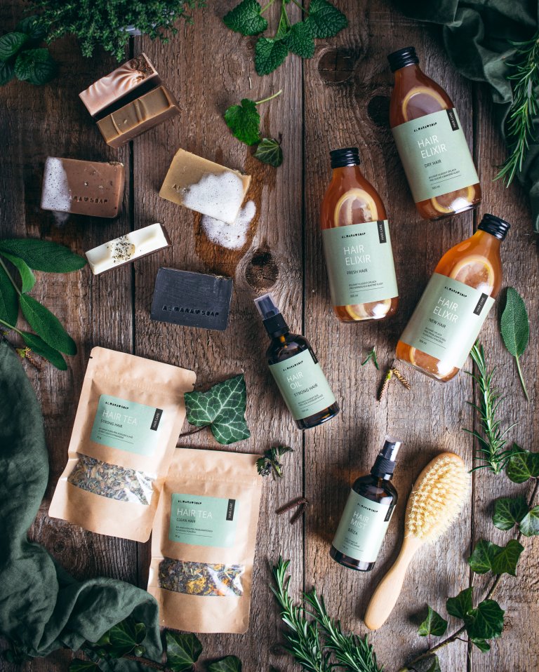 Upper view photo of Almara Soap hair care products with green labels on a dark brown wooden background with leaves of various herbs that these products contain.