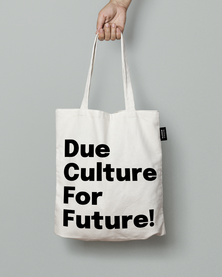 White canvas bag with claim Due Culture For Future in black, Nuraga Budaya logo in white is placed on the black label on the right side of the bag. Sample merchandise for Nuraga budaya.