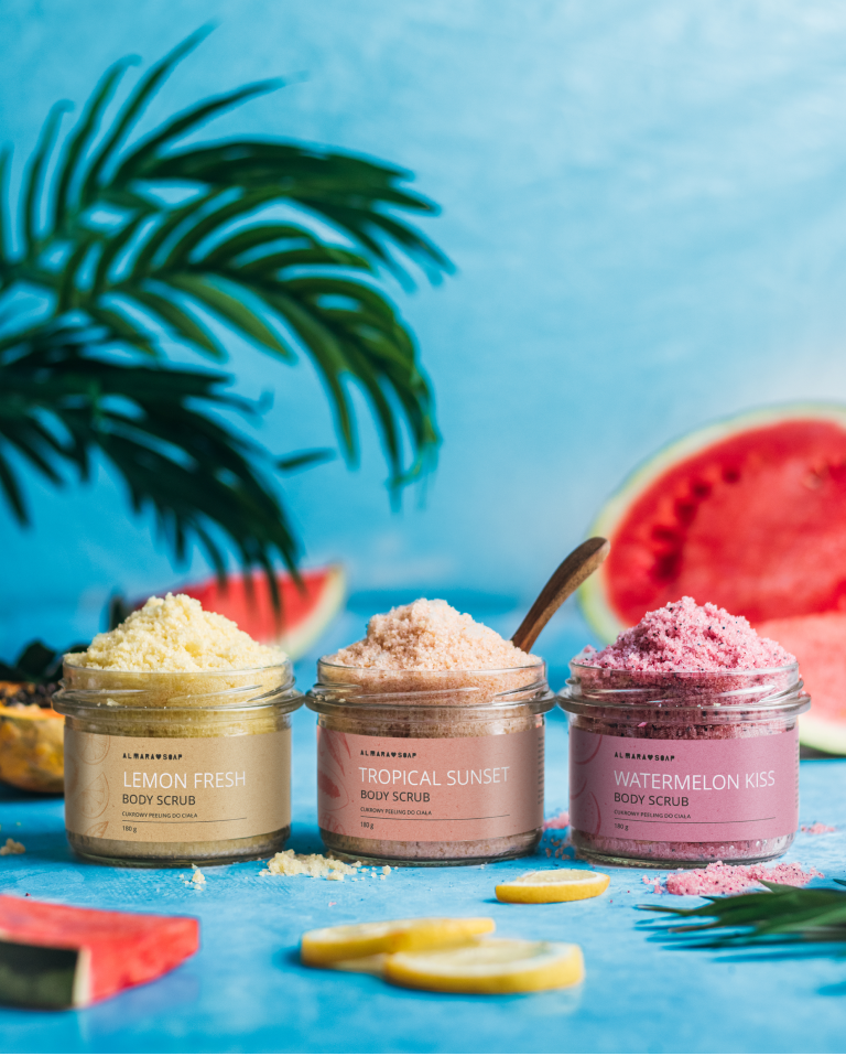 Photo of three fruit-scented body scrubs from the summer limited edition on a turquoise background with palm leaves, watermelon and lemon slices as decoration. Exotic mood.
